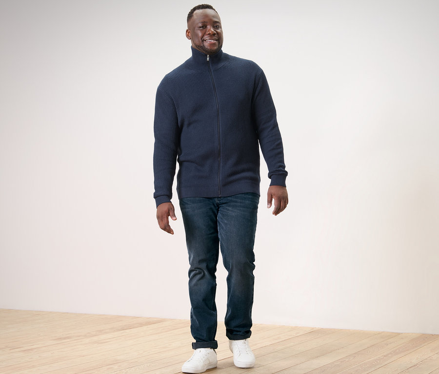 Un homme marche et porte une jaquette en maille bleu marine, un jean et des baskets.