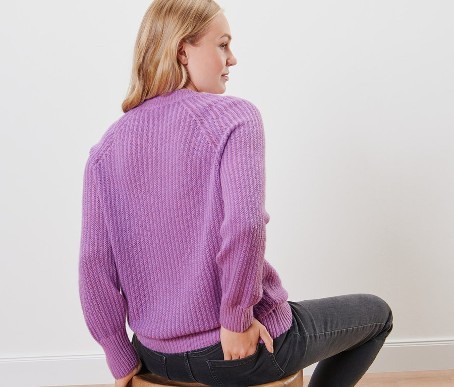 Une femme est assise sur un tabouret, dos à l'appareil photo, et porte un pull en maille lilas.