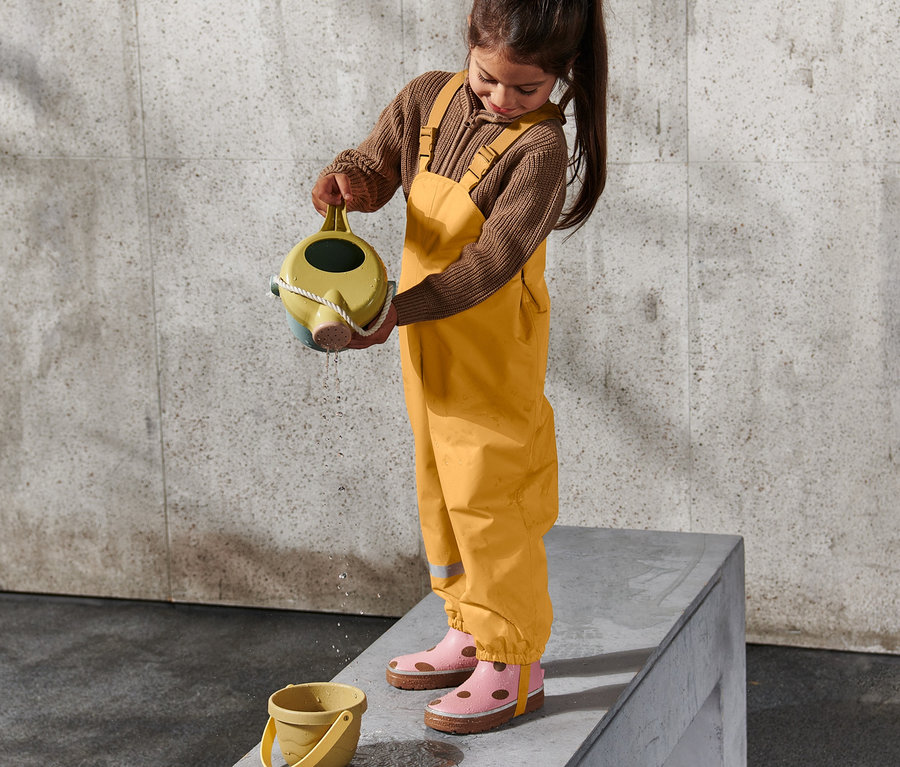 Une fille en pantalon de pluie thermique pour enfant, jaune, verse de l'eau d'un arrosoir dans un seau jaune.