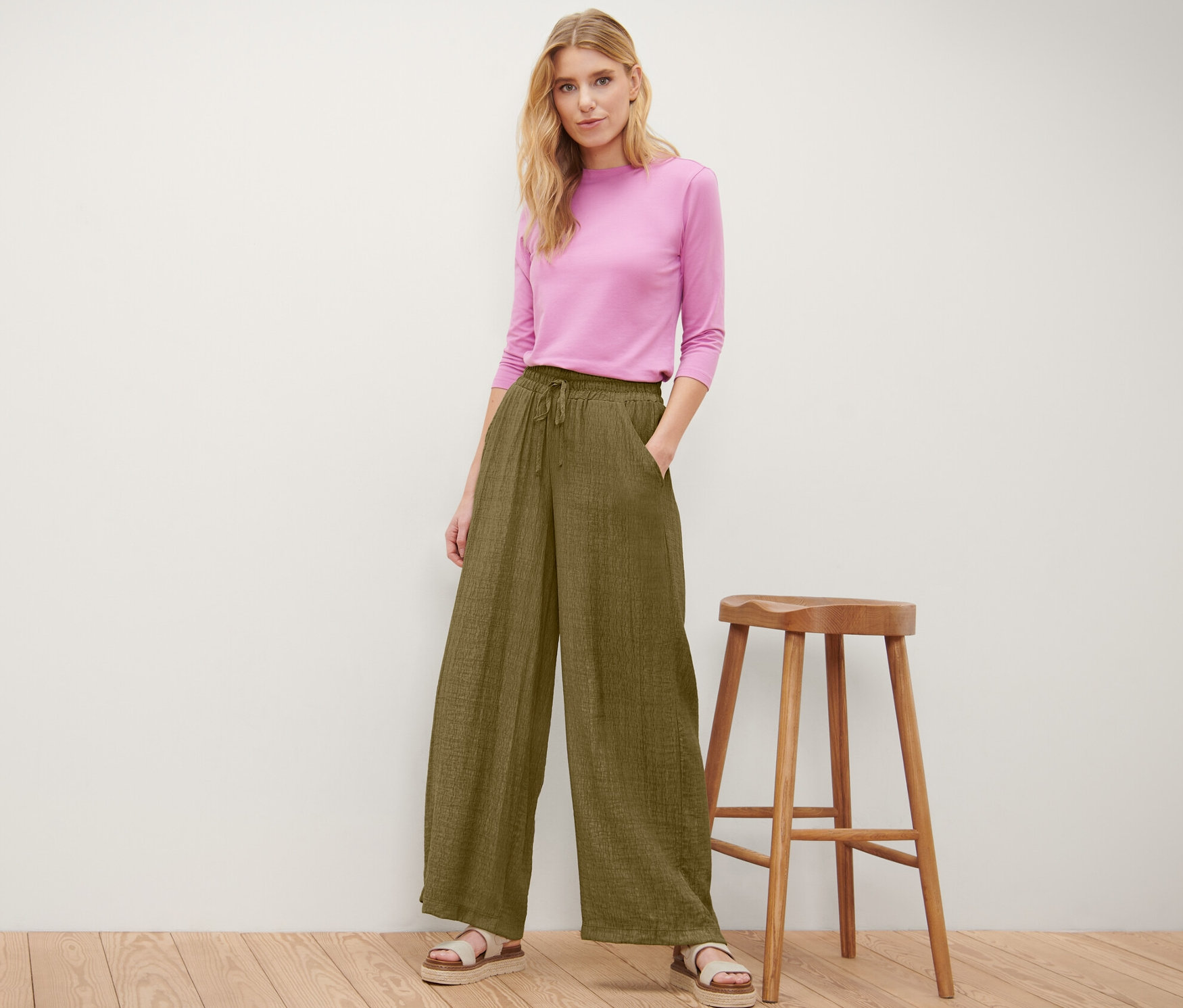 Une femme pose en pantalon à taille élastique olive et haut rose à côté d'un tabouret en bois.