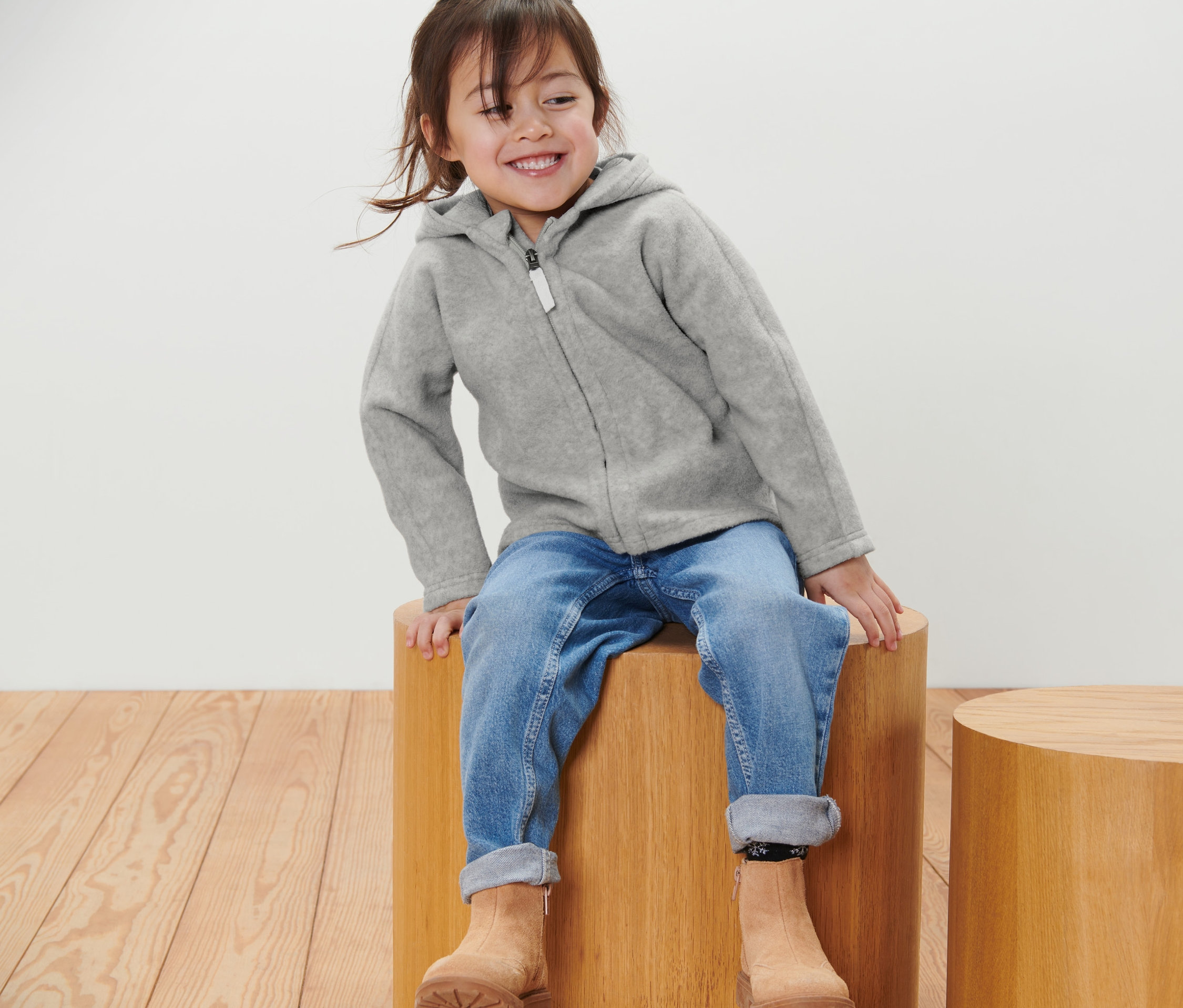 Fillette souriante assise sur un tabouret en bois portant une veste polaire pour enfant grise, un jean et des bottes marron.