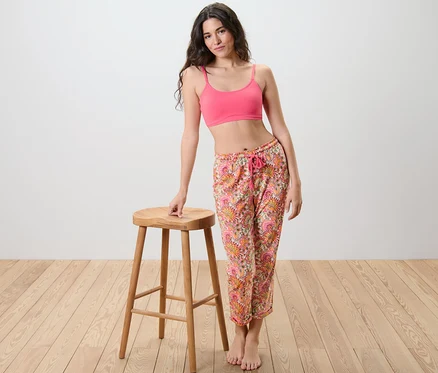 Une femme pose à côté d'un tabouret, portant un haut rose et un pantalon de pyjama.