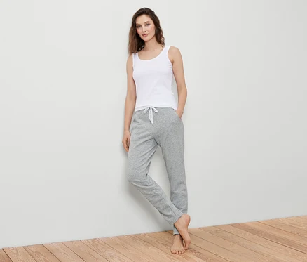 Une femme en débardeur blanc et pantalon de survêtement gris se tient contre un mur.