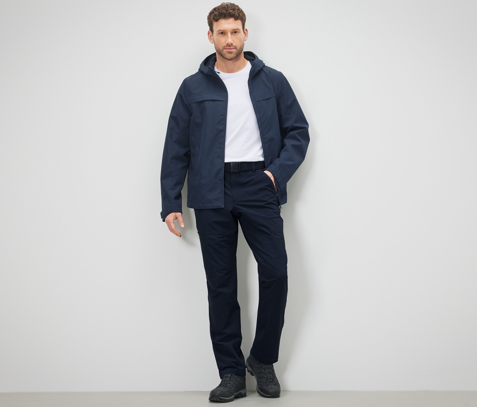 Un homme pose en pantalon et veste d'extérieur devant un mur.