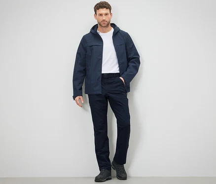 Un homme pose en pantalon et veste d'extérieur devant un mur.