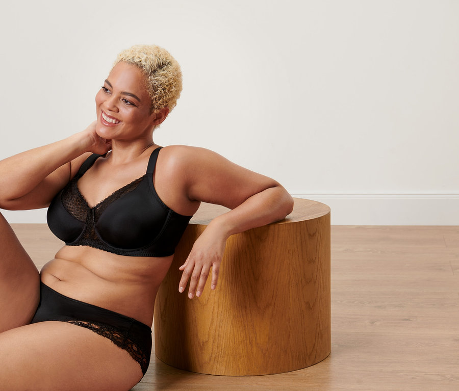 Une femme est assise sur le sol, appuyée contre un tabouret en bois, elle porte un soutien-gorge minimiseur noir.