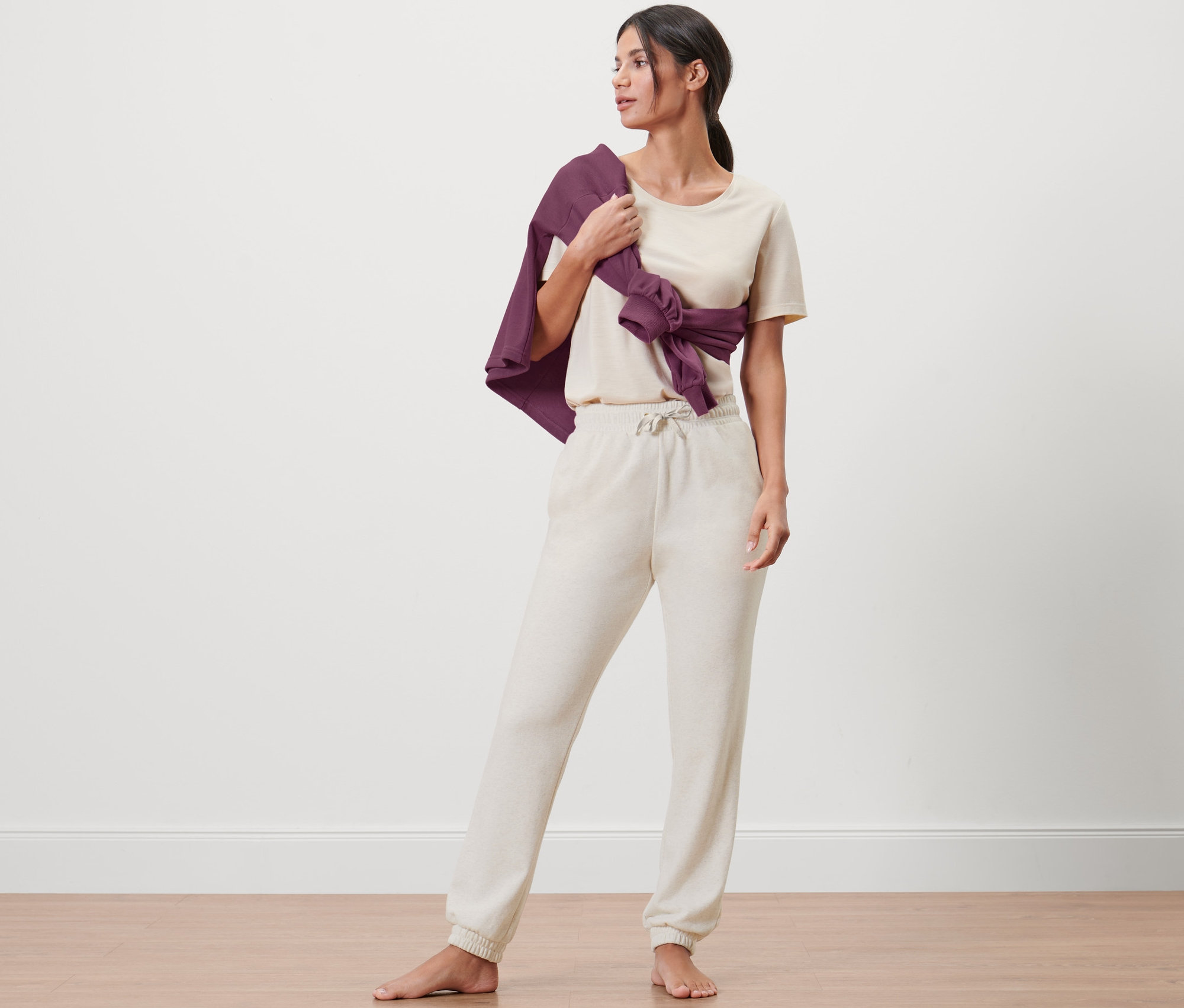 Une femme se tient pieds nus en pantalon de survêtement beige et T-shirt. Un cardigan violet est noué autour de sa taille.