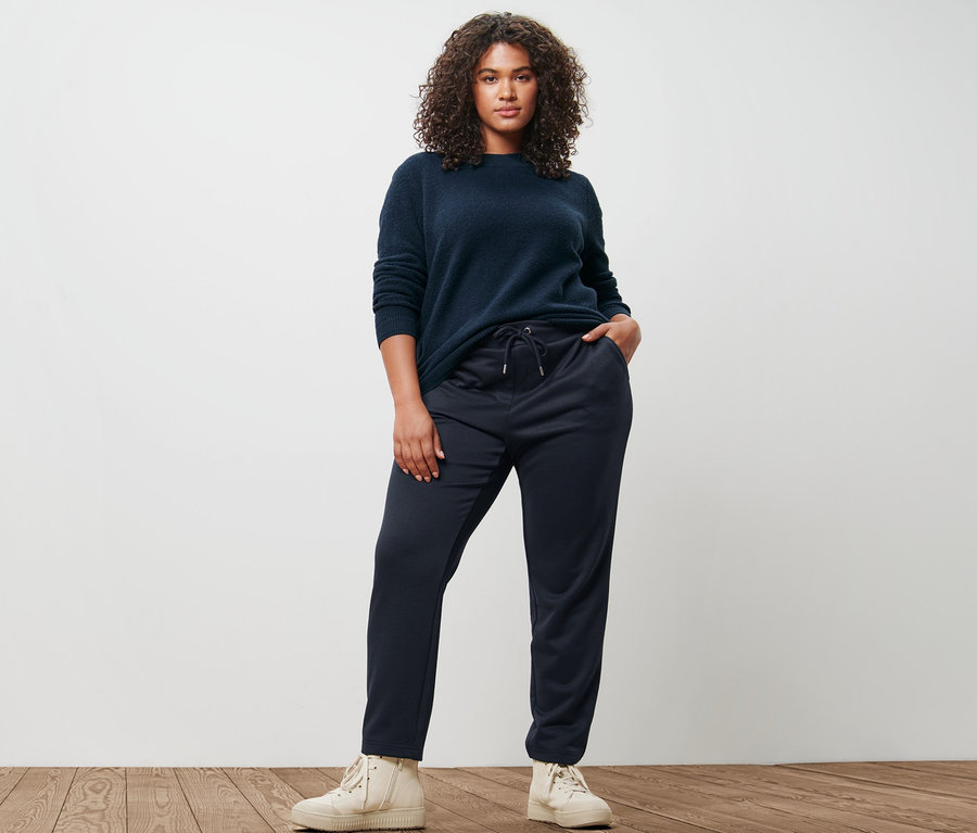 Une femme pose, portant un pull en maille aspect bouclé et un jogpant bleu foncé.