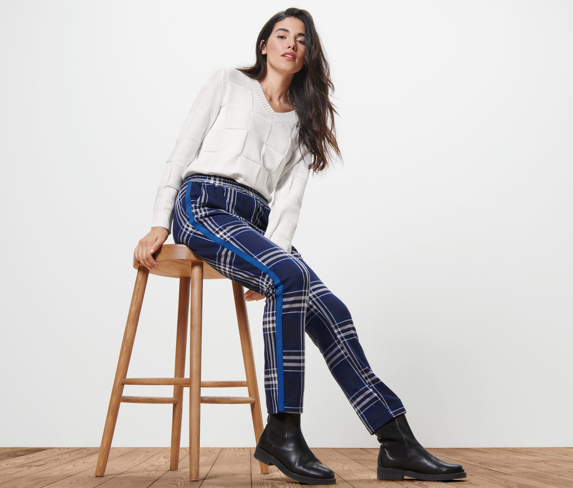 Femme assise sur un tabouret en bois portant un pantalon de jogging à carreaux et un pull.