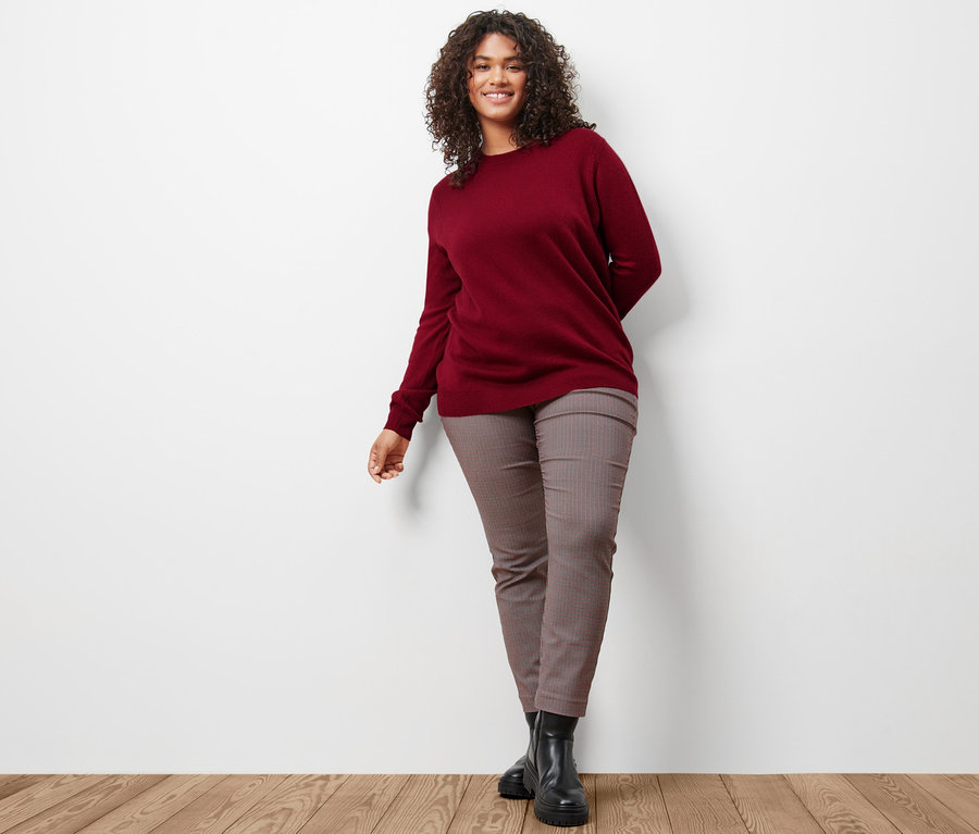 Une femme porte un pull en maille fine rouge bordeaux avec du cachemire et un pantalon stretch à carreaux.