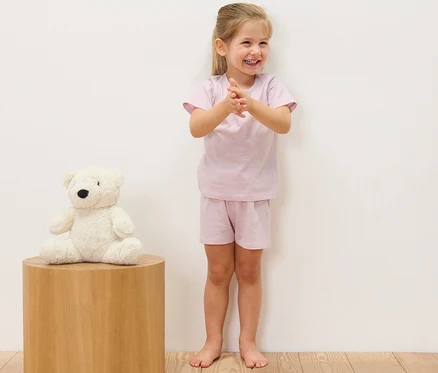 Une fillette souriante applaudit, vêtue d'un pyjama court rayé pour enfant (lot de 2). Un ours en peluche est assis à gauche sur un tabouret en bois.