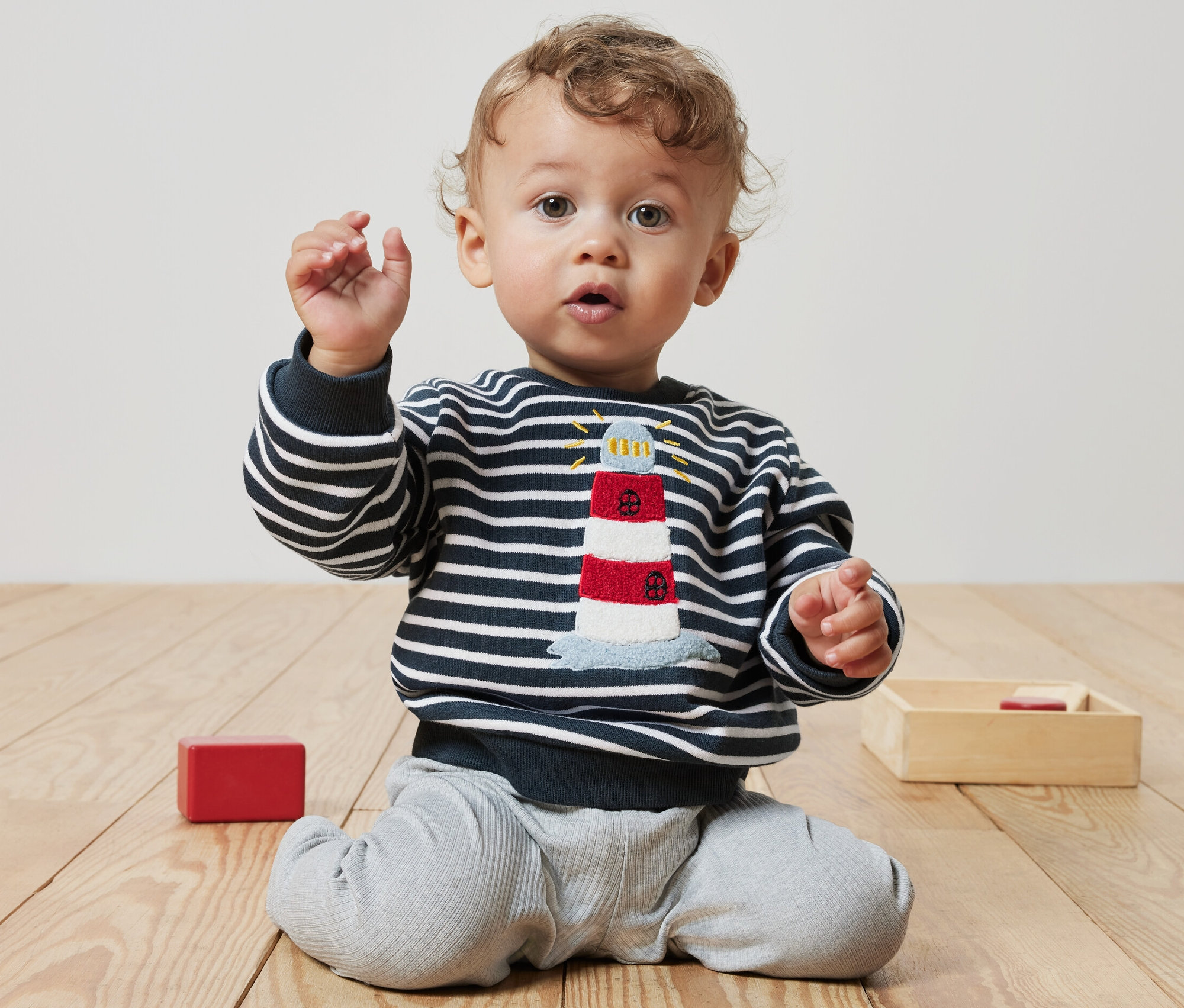 Un bébé bouclé est assis par terre et porte un sweatshirt pour bébé avec un motif de phare.