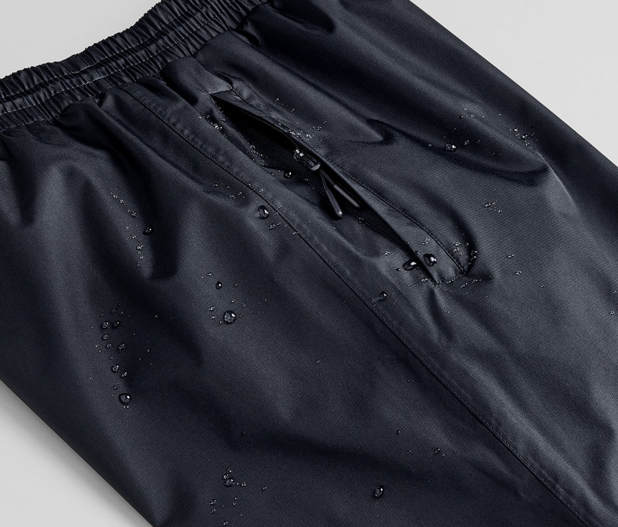 Gros plan d'un pantalon de pluie unisexe noir avec des gouttes d'eau et une poche zippée.