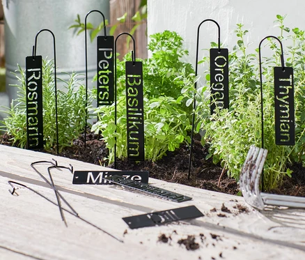 Des écriteaux noirs pour herbes sont piqués dans un parterre d'herbes, des outils de jardinage sont posés sur une table en bois.