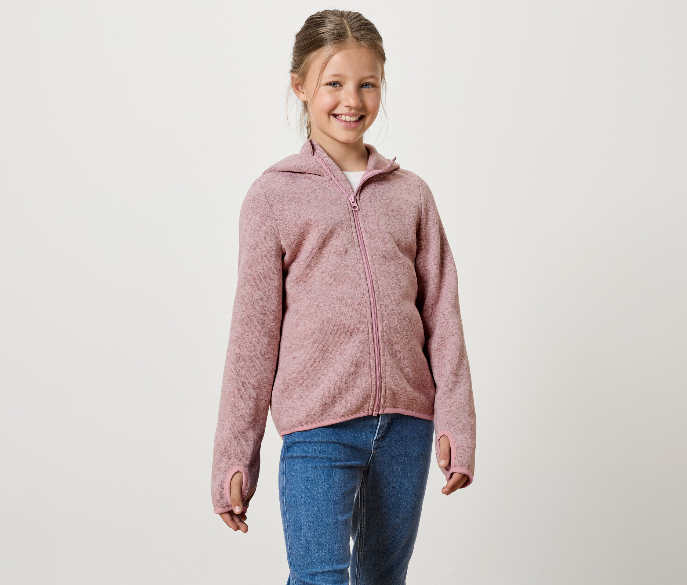 Une fille souriante porte un sweat à capuche rose zippé et un jean bleu.