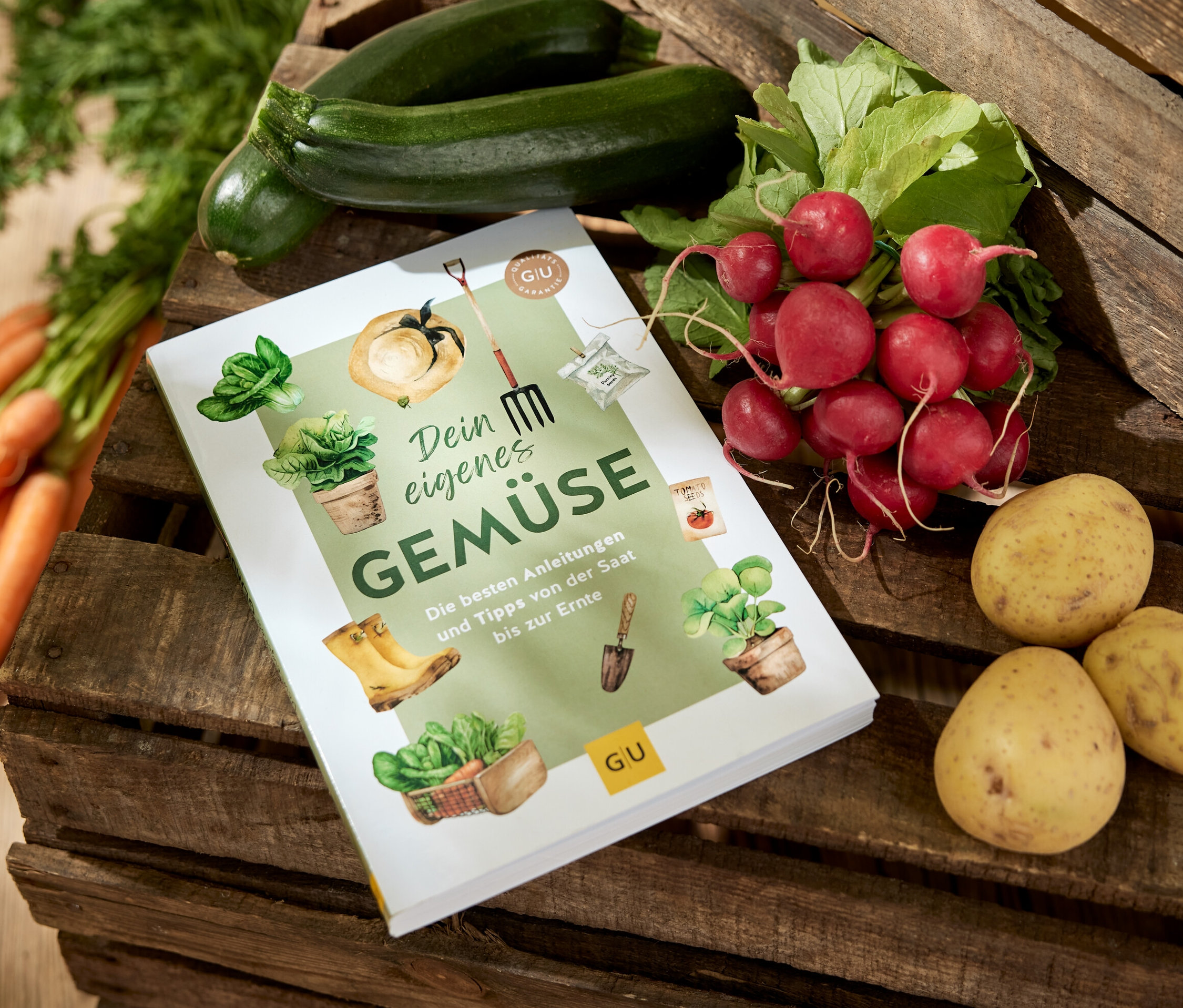 Le livre « Dein eigenes Gemüse » (tes propres légumes, en allemand) est posé sur une caisse en bois, entouré de carottes, de courgettes, de radis et de pommes de terre.