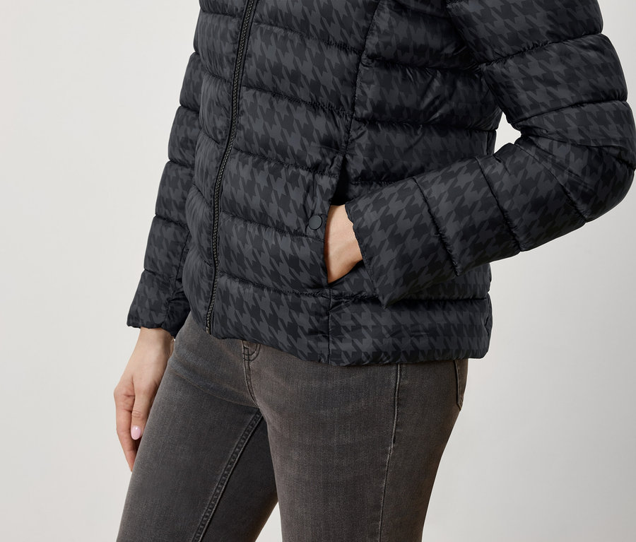 Une femme a la main dans la poche d'une doudoune noire à motif pied-de-poule et d'un jean.