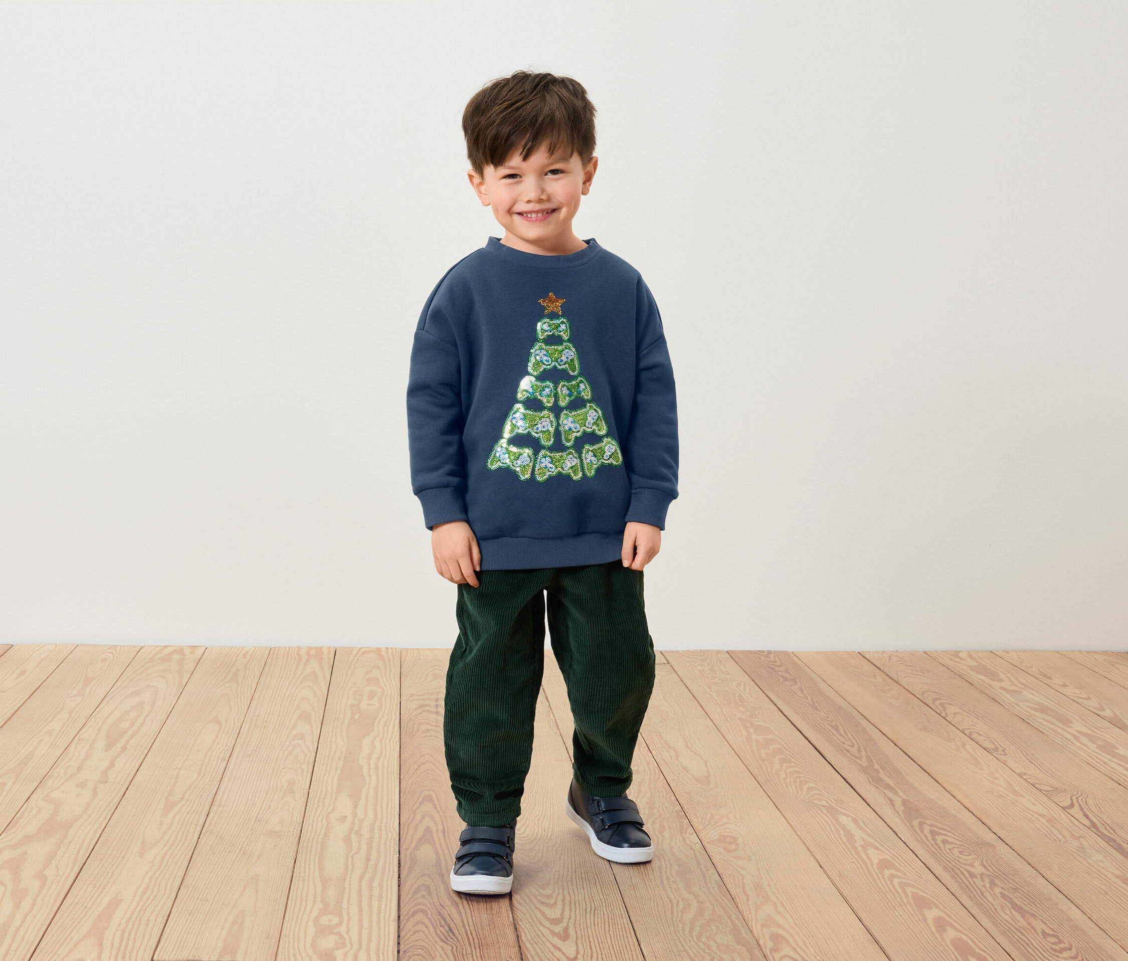 Un garçon souriant debout porte un pull bleu marine avec un motif de sapin de Noël fait de manettes de jeu, un pantalon vert et des chaussures bleues.