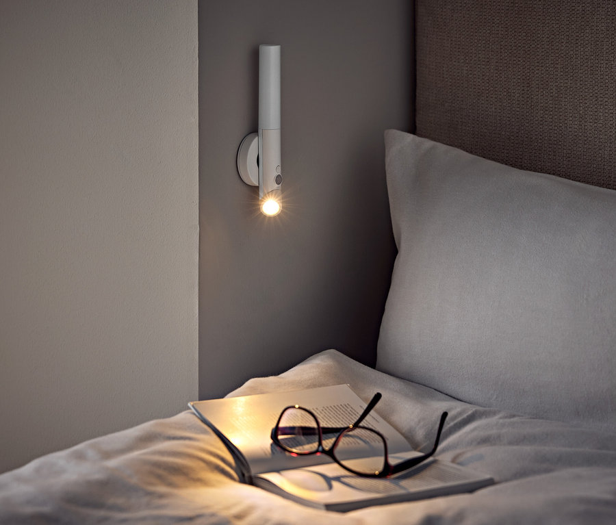 Sur un lit, des lunettes et un livre ouvert sous une applique murale.