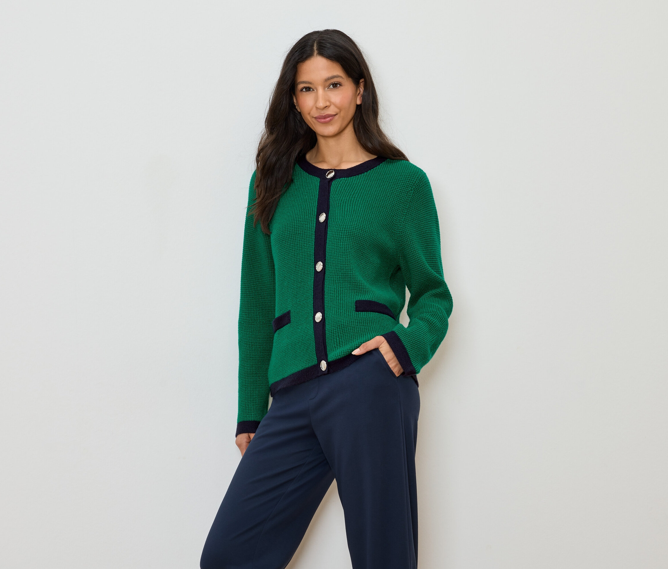 Une femme aux cheveux foncés porte un cardigan vert avec des détails bleu marine et un pantalon bleu marine.
