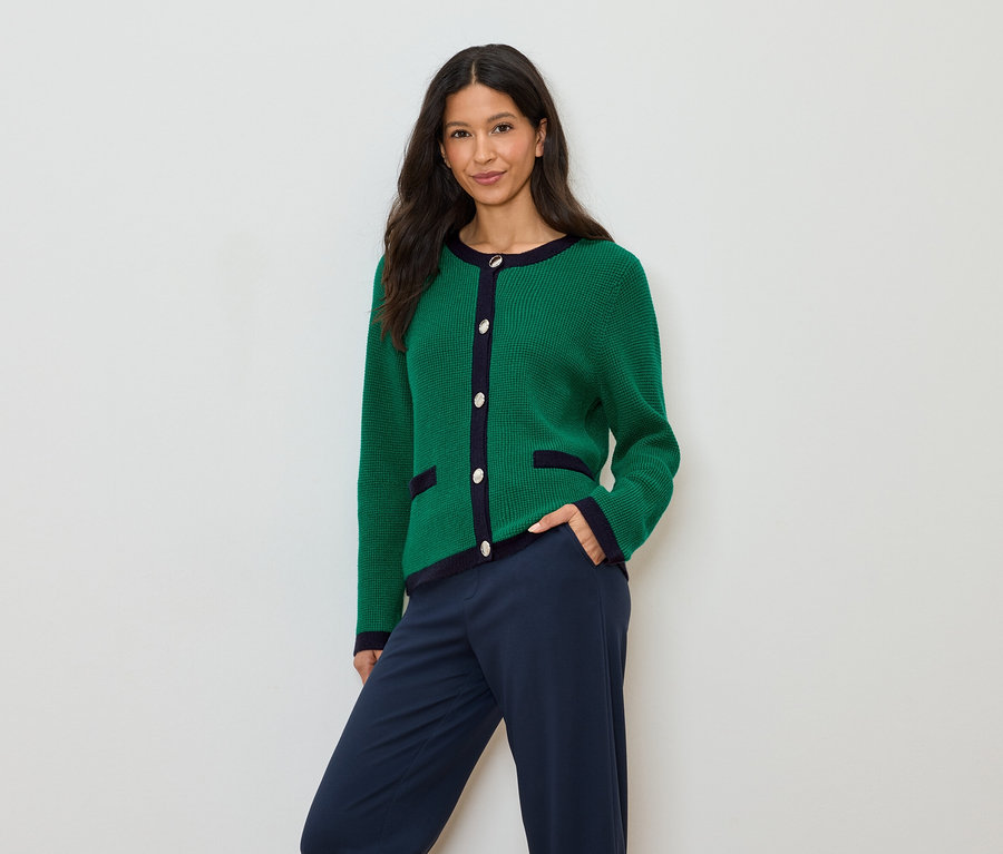 Une femme aux cheveux foncés porte un cardigan vert avec des détails bleu marine et un pantalon bleu marine.
