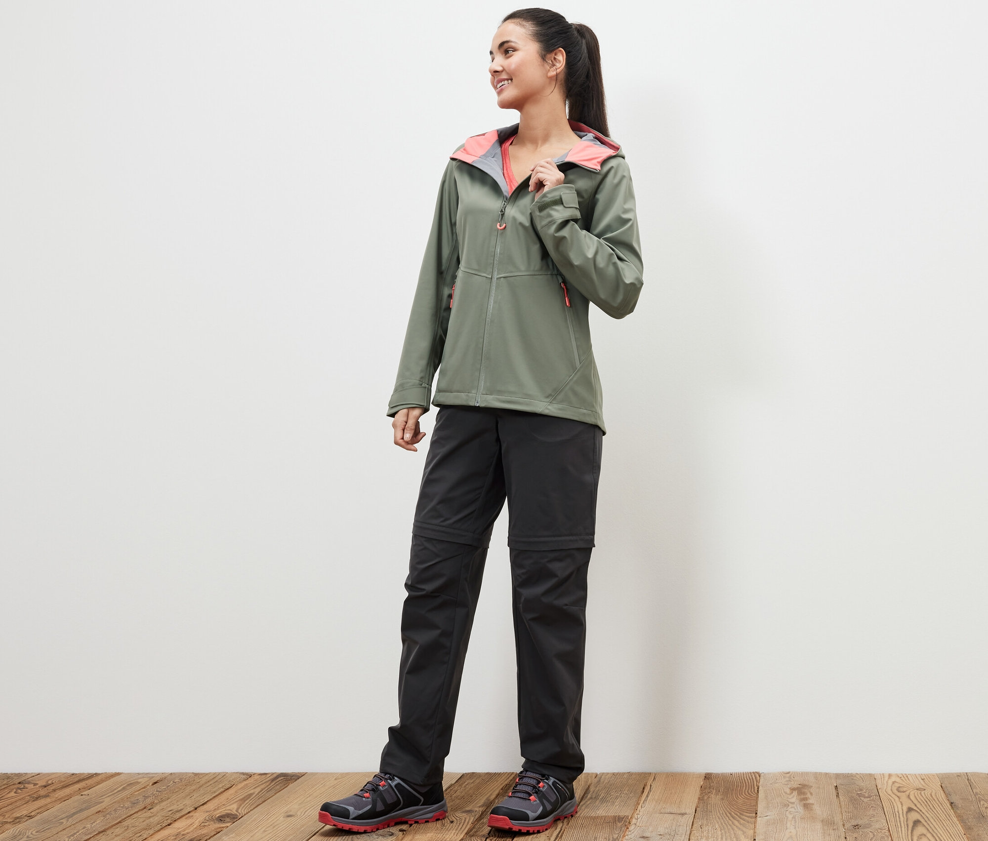Une femme debout sur un plancher en bois porte une veste verte et un pantalon technique 2 en 1 noir.