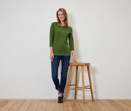 Une femme portant un t-shirt olive à manches trois-quarts et un jean bleu foncé se tient debout, appuyée sur un tabouret en bois.