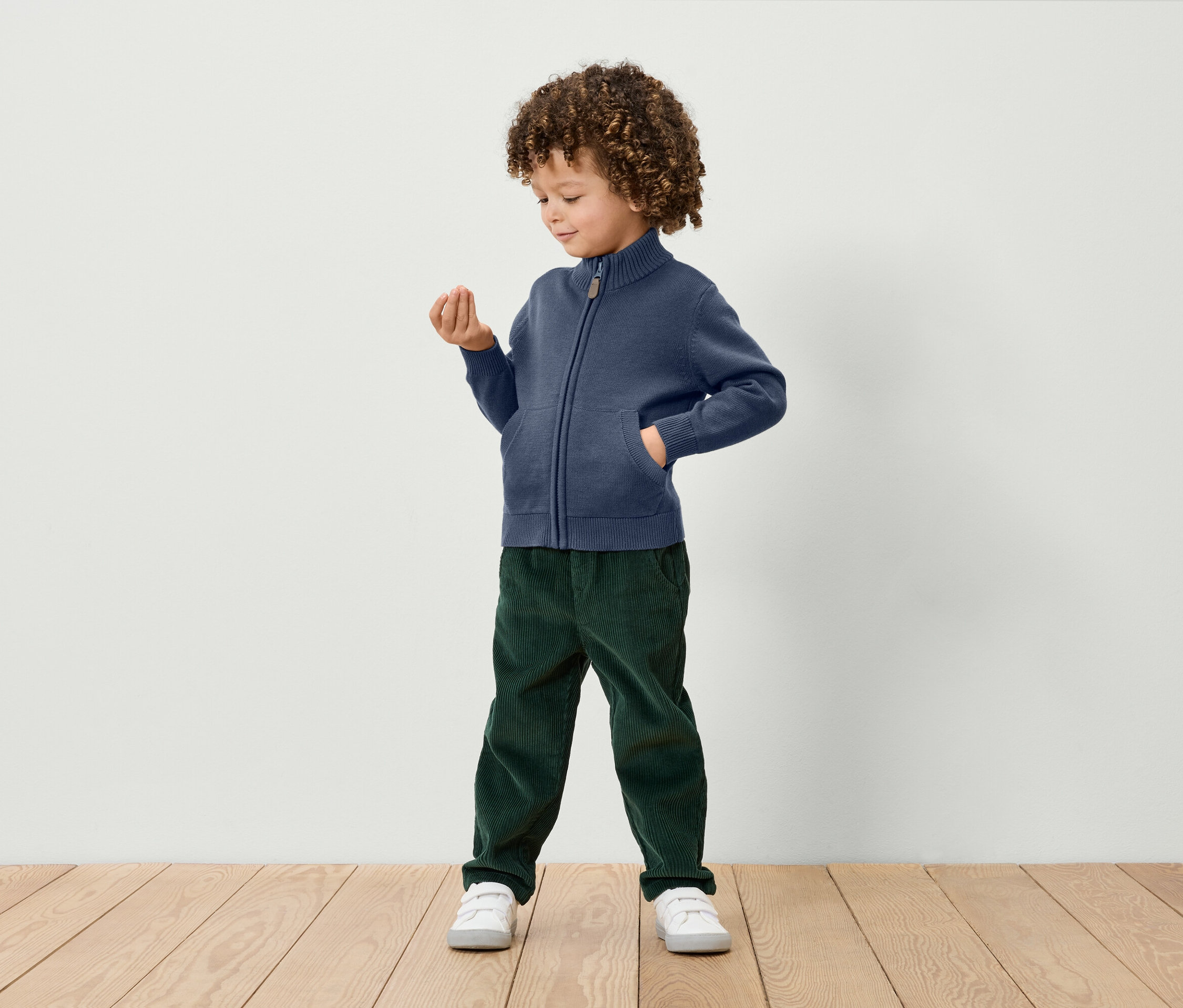 Un garçon aux cheveux bouclés se tient debout et tient un petit objet dans sa main. Il porte un cardigan bleu zippé, un pantalon de velours vert et des baskets blanches.