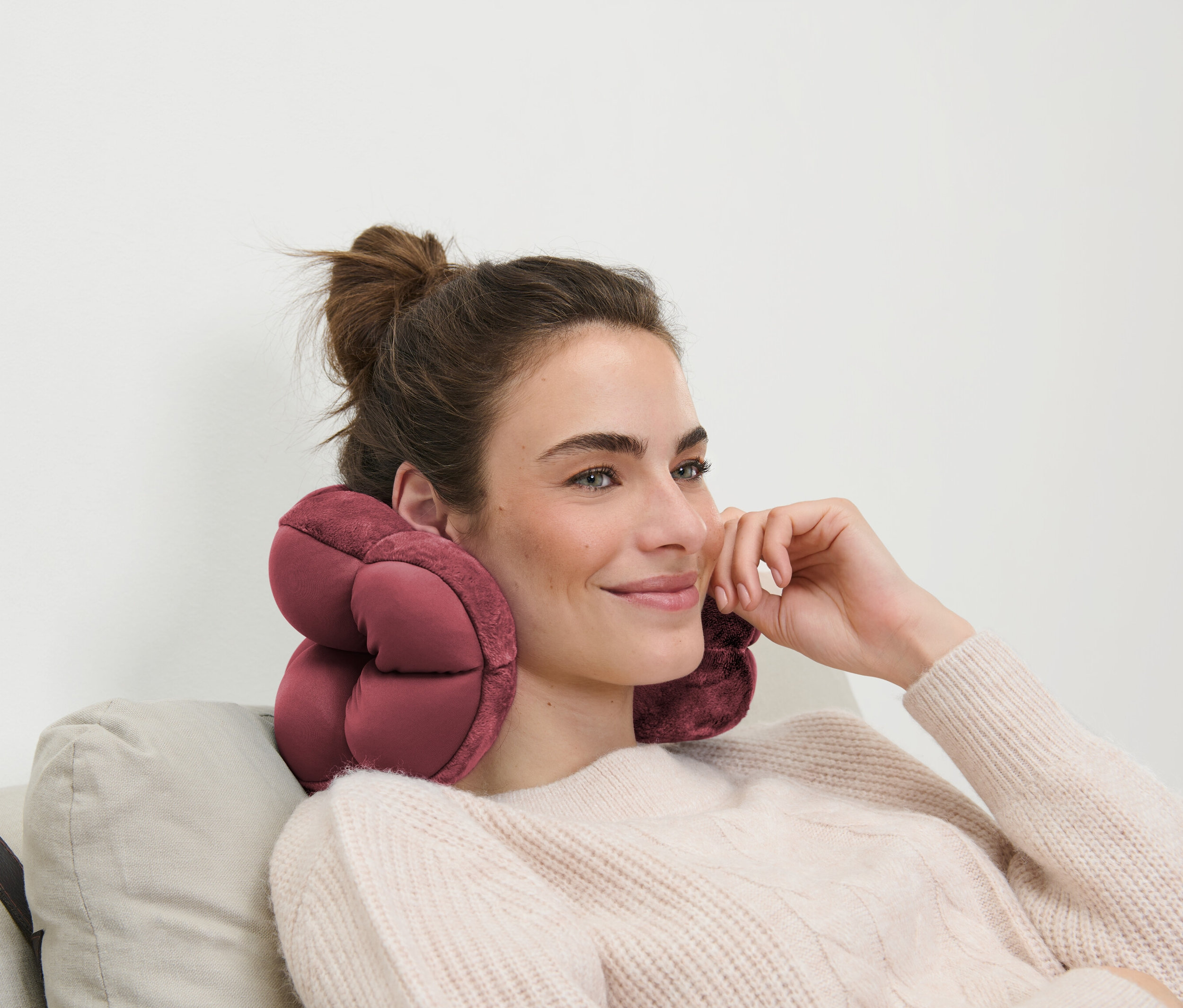 Une femme aux cheveux bruns est assise sur un canapé avec un oreiller de voyage rose autour du cou.