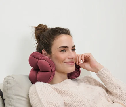 Une femme aux cheveux bruns est assise sur un canapé avec un oreiller de voyage rose autour du cou.