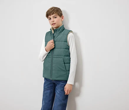 Un garçon ferme une veste matelassée verte sur un T-shirt à manches longues blanc et un jean bleu.