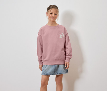 Une fille porte un sweat-shirt rose avec un logo LA et une jupe en jean.