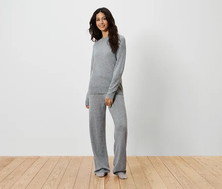 Une femme se tient sur un plancher en bois et porte un pull et un pantalon en tricot gris.
