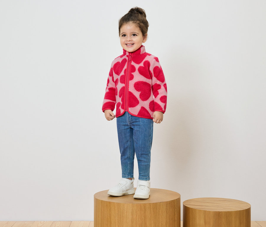 Une petite fille souriante se tient sur un socle en bois et porte une veste polaire rose avec un motif de coeurs rouges, un jean et des baskets blanches.