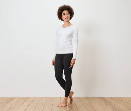 Une femme debout porte un t-shirt blanc à manches longues et un legging noir.
