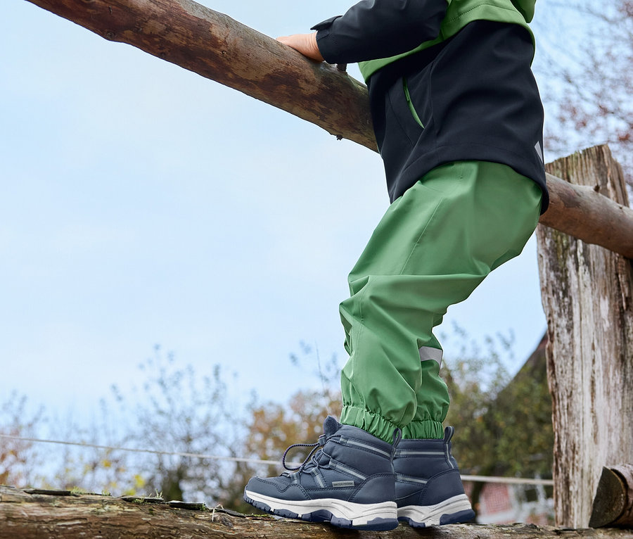 Un garçon grimpe sur une clôture en bois. Il porte un pantalon de pluie pour enfant vert gazon, des bottines thermiques pour enfant vertes et une veste en softshell pour enfant.