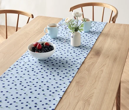 Sur une table en bois, un chemin de table à motif de myrtilles est disposé avec un bol de fruits, deux tasses bleues et un vase de fleurs.