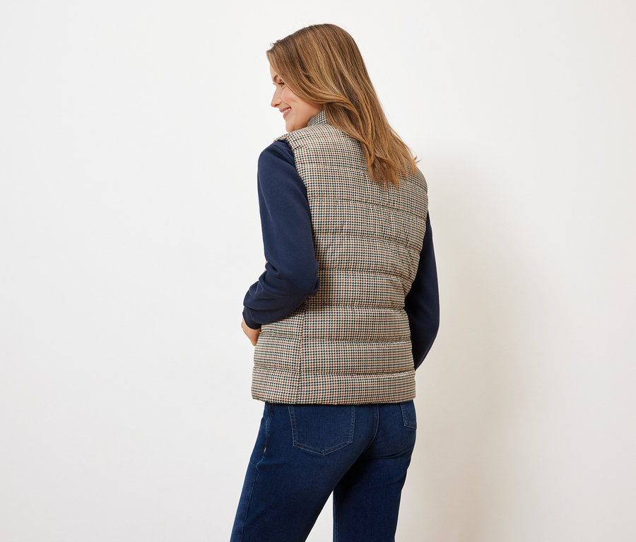 Une femme, dos à la caméra, porte un gilet matelassé pied-de-poule, un pull bleu et un jean.