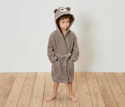 Un enfant aux cheveux bouclés se tient sur un plancher en bois, portant un peignoir marron à capuche avec des oreilles d'ours.