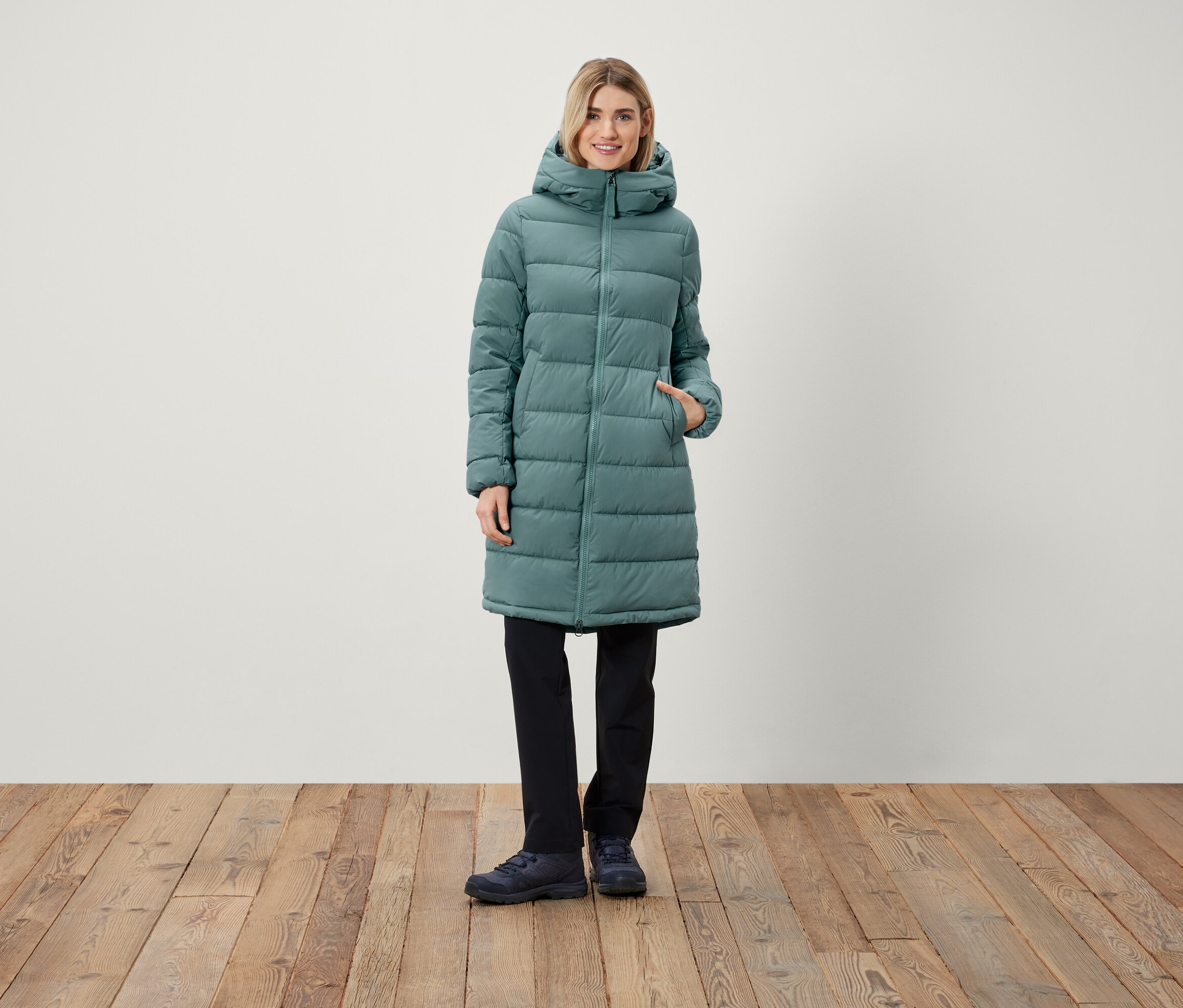 Une femme debout porte un long manteau matelassé à capuche, un pantalon noir et des chaussures.