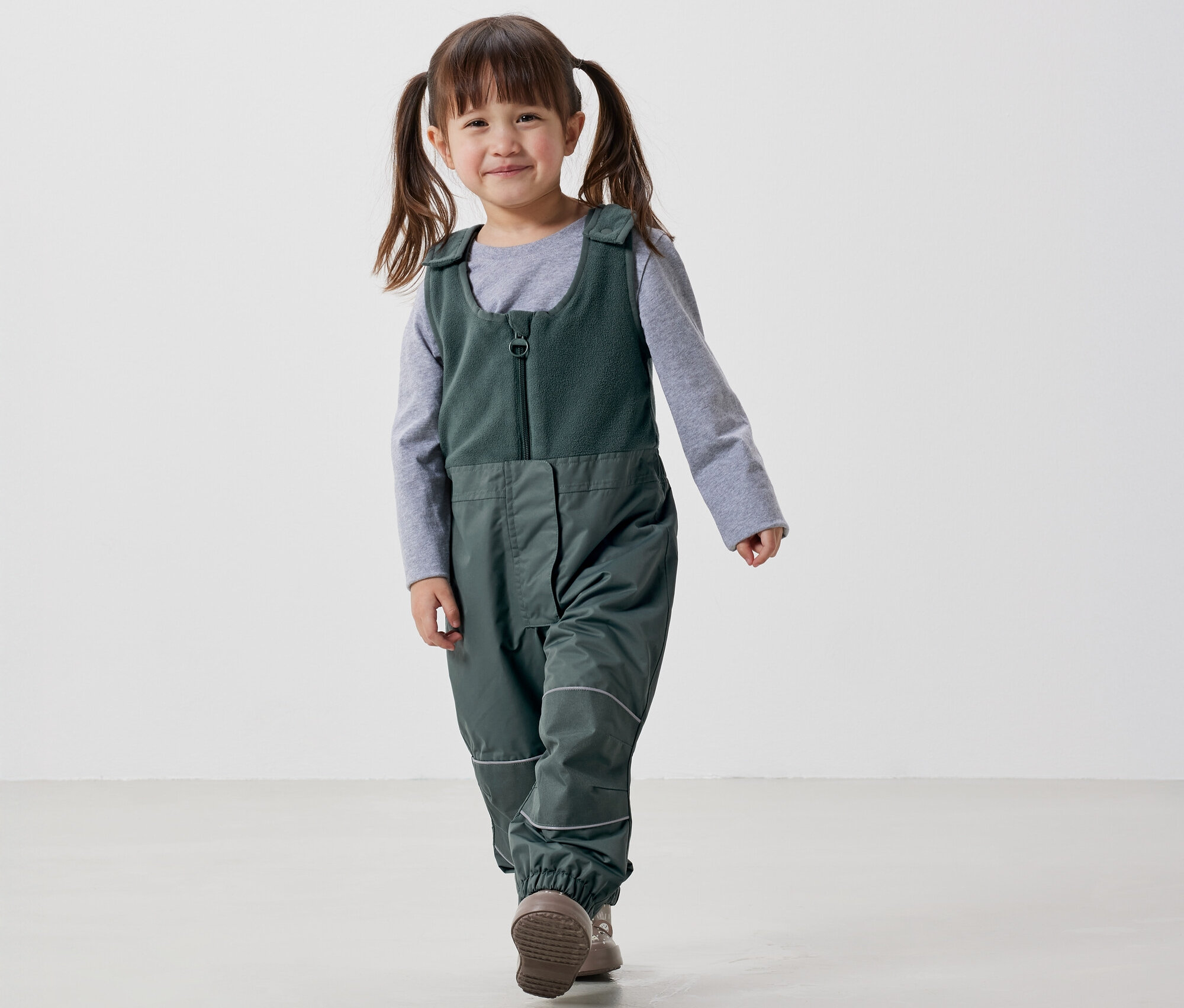 Fillette souriante avec des couettes marchant dans une salopette verte et un t-shirt gris sur fond blanc.