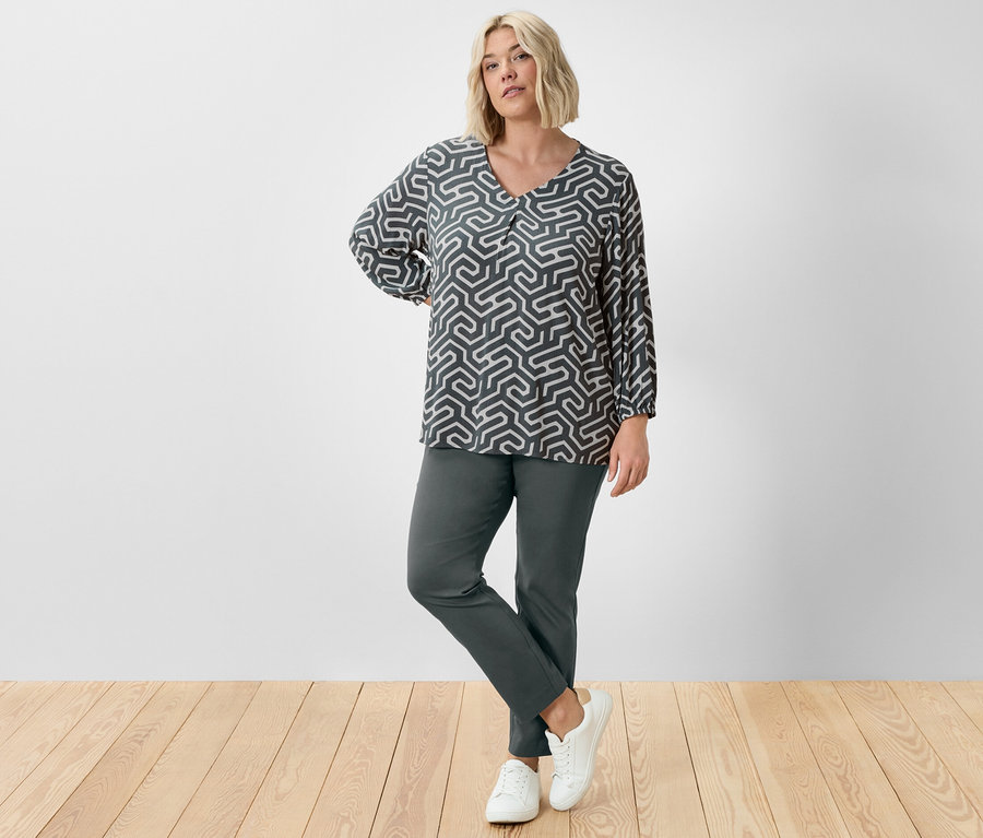Une femme pose en studio en pantalon stretch vert foncé et blouse aspect froissé.