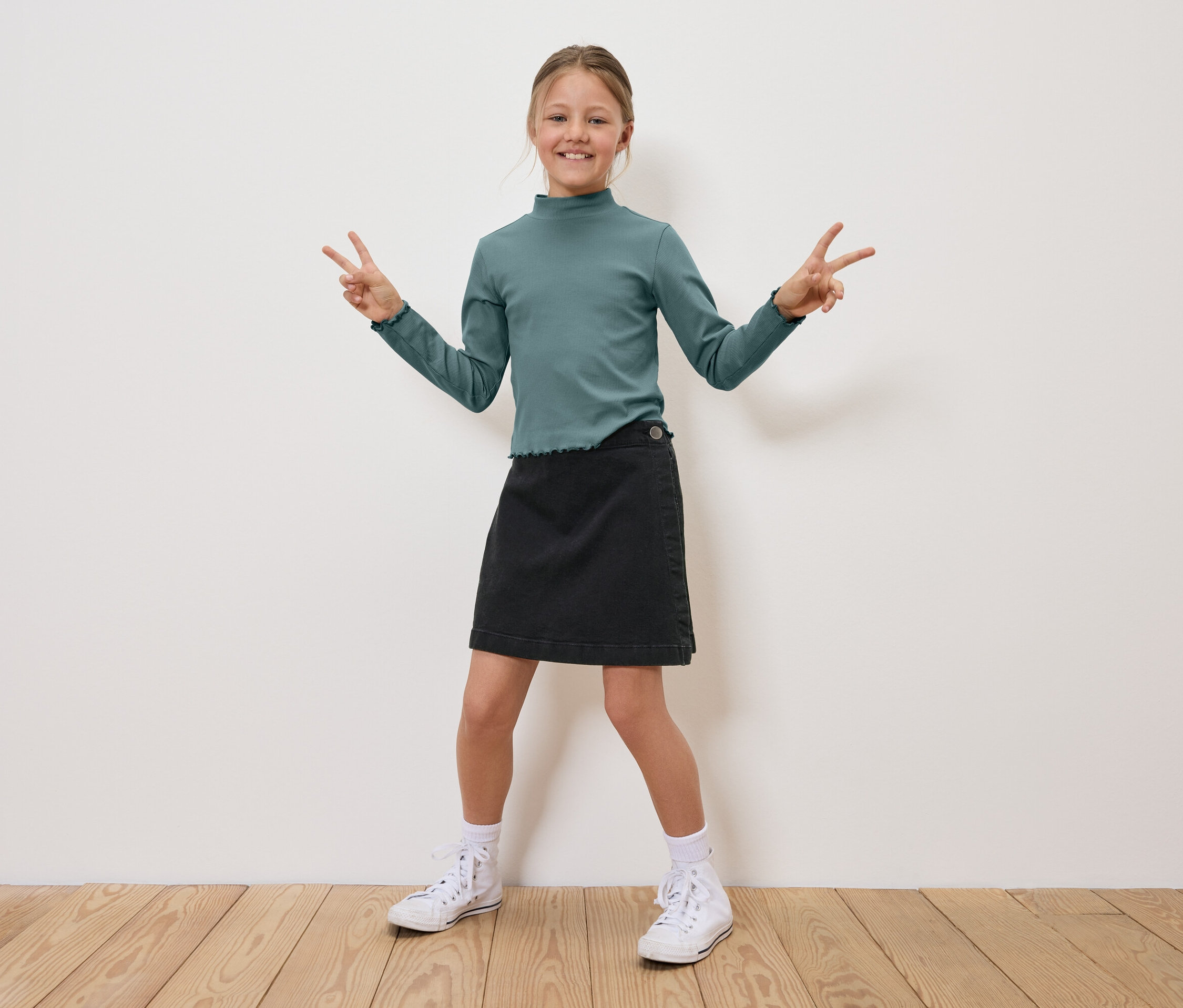 Une fille portant un haut vert et une jupe noire se tient debout et fait le signe de la victoire avec ses doigts.