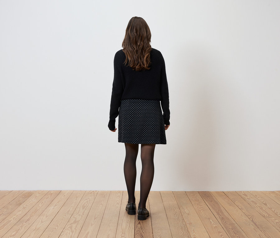 Une femme se tient dos à la caméra, portant un pull noir, une jupe à motifs et des collants noirs.