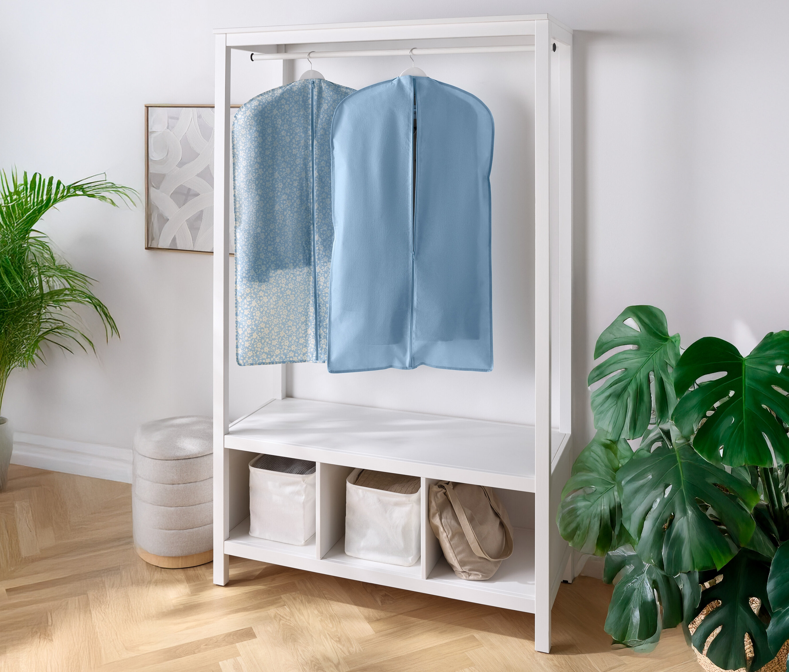 Deux housses à vêtements suspendues à une armoire blanche, avec des étagères et des compartiments en dessous. Une plante d'intérieur se trouve à gauche et à droite.