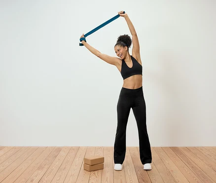 Une femme souriante s'étire avec une bande de résistance bleue au-dessus de sa tête. Deux blocs de yoga sont posés sur le sol.