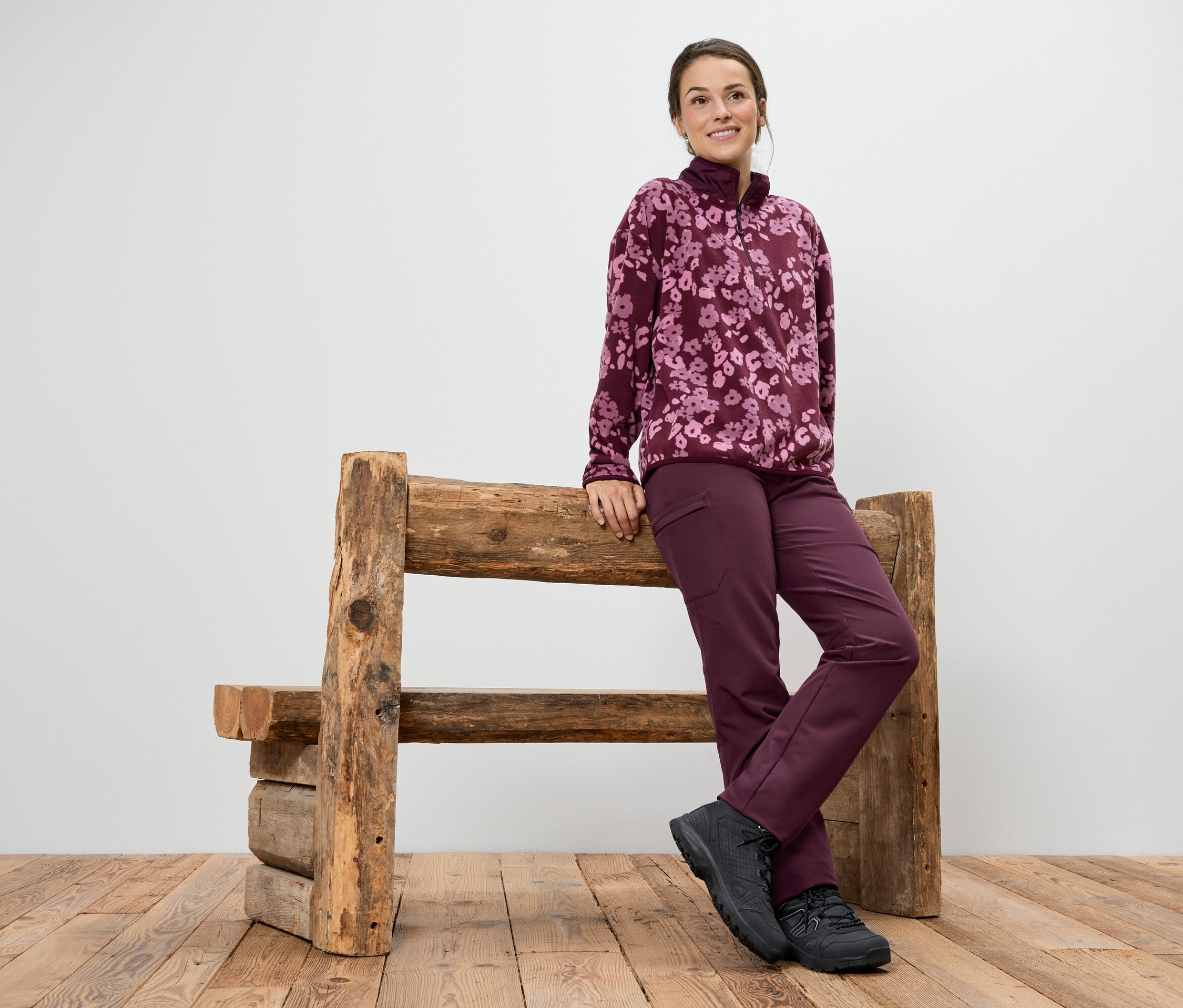Une femme portant un pull en polaire violet à motif floral et un pantalon est appuyée contre un banc en bois.