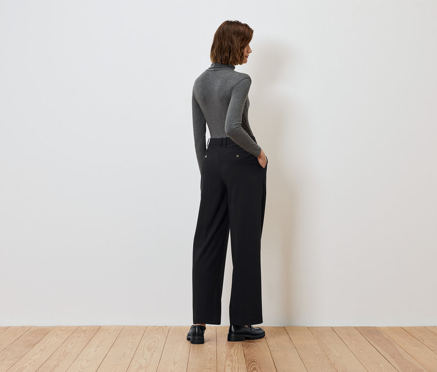 Vue arrière d'une femme avec les mains dans les poches d'un pantalon noir et un col roulé gris.