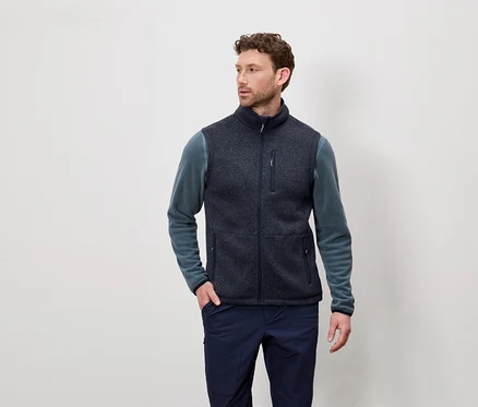 Un homme aux cheveux courts et bouclés porte un gilet en polaire bleu marine et un T-shirt à manches longues bleu-vert et pose devant un fond clair.