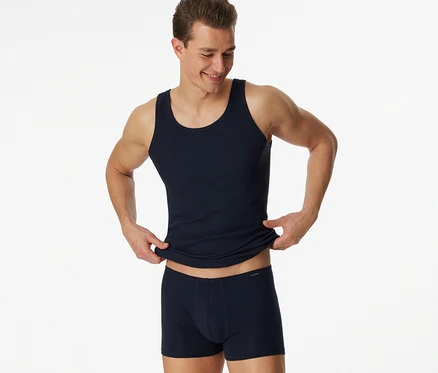 Homme souriant portant un maillot de corps SCHIESSER bleu en maille côtelée fine et un boxer bleu, remontant le maillot de corps des deux mains.